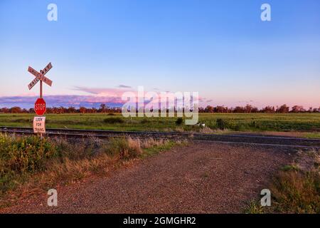 Eisenbahnüberquerung der nicht versiegelten Schotterstraße auf Bauernhöfen in der Nähe der ländlichen australischen Stadt Moree auf einem künstlerischen Becken - Sonnenuntergangslandschaft. Stockfoto