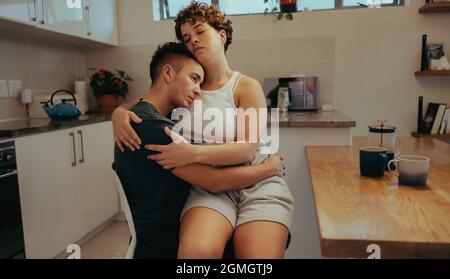 Junge Frau, die sich zu Hause mit ihrem Freund verklebt. Anhänglich junge Frau umarmt ihren Freund, während sie auf seinem Schoß sitzt. Romantisches junges queeres Paar Stockfoto