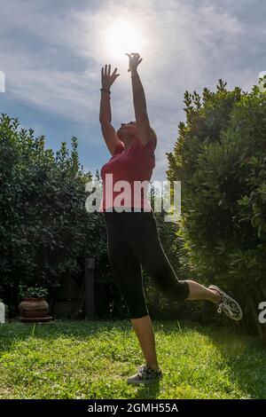 Eine weiße Frau mittleren Alters gibt vor, die Sonne zu ergreifen, während sie auf einem Bein balanciert Stockfoto