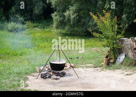 Alter gusseiserner Kessel auf einem Feuer. Kochen von Speisen im Freien in einem Topf auf einem Stativ. Axt in einem Holzstumpf. Mittelalterlicher Ritter Schild und Kupferpfanne Stockfoto