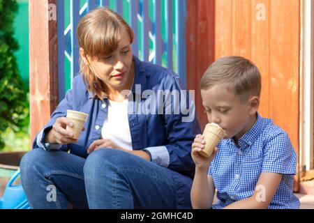 Vorschulkinder Junge mit Mama essen Eis auf der Veranda eines Hauses im Dorf an einem sonnigen Sommertag. Selektiver Fokus. Hochformat Stockfoto