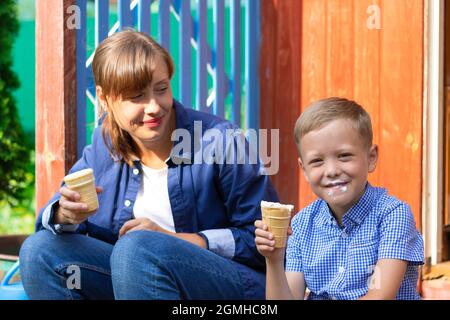 Vorschulkinder Junge mit Mama essen Eis auf der Veranda eines Hauses im Dorf an einem sonnigen Sommertag. Selektiver Fokus. Hochformat Stockfoto