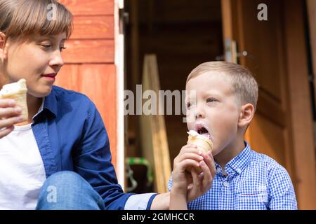 Vorschulkinder Junge mit Mama essen Eis auf der Veranda eines Hauses im Dorf an einem sonnigen Sommertag. Selektiver Fokus. Hochformat Stockfoto