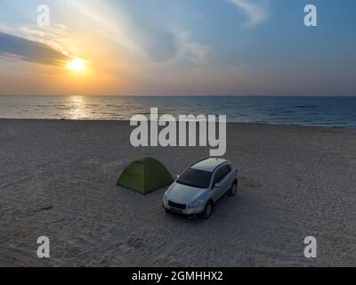 Sonnenaufgang über dem Zelt, das am Strand steht, dem Sandstrand und Touristen, die sich in einem Zelt neben dem Auto ausruhen Stockfoto
