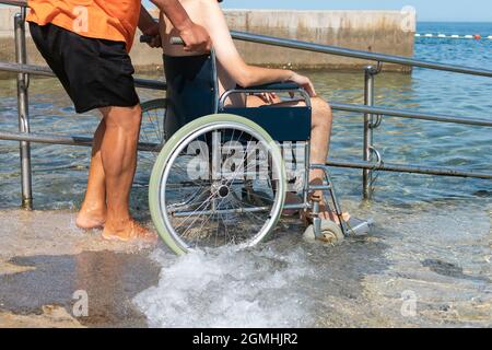 Mann schiebt langsam und vorsichtig behinderte Person, die in einem Rollstuhl sitzt, entlang der Rampe am Strand ins Meer Stockfoto