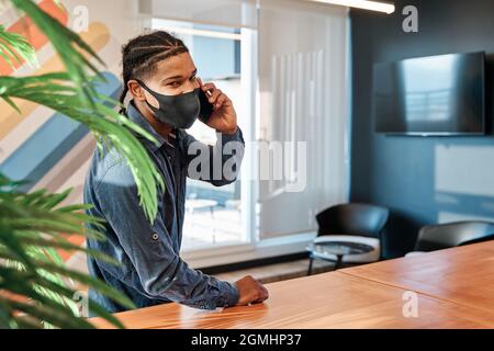 Junger latein, der sich auf einen Tisch lehnt und mitten in der Kuhskräone mit einer Maske und in Quarantäne, sozialem Distanzierungskonzept telefoniert Stockfoto