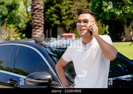 Ein junger asiatischer Geschäftsmann, ein Mann in einem weißen Hemd und einer Sonnenbrille, steht in der Nähe eines modernen Autos und spricht auf einem Mobiltelefon, löst Probleme und Fragen, Stockfoto