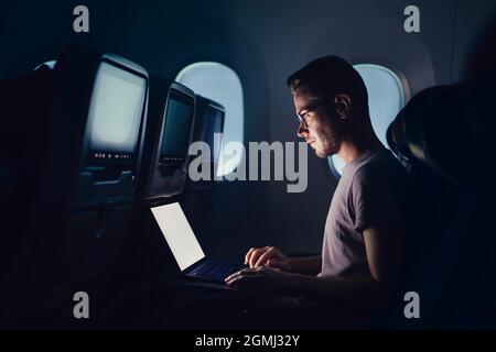 Mann, der mit dem Flugzeug reist. Junger Passagier, der während des Nachtflugs einen Laptop benutzt. Stockfoto
