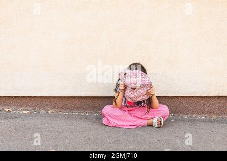 Das kleine Brünette Mädchen versteckt sich hinter ihrem Rucksack, sitzt allein und fühlt sich traurig Stockfoto