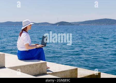 Arbeiten Sie von überall aus. Seitenansicht einer jungen Frau, Freiberuflerin im Strohhut, die am Laptop arbeitet, während sie am Strand sitzt Stockfoto
