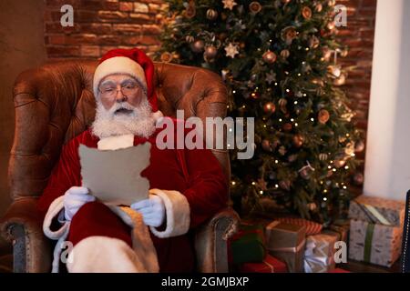 Ernsthafter weißbärtiger Weihnachtsmann, der in einem gemütlichen Sessel gegen den Weihnachtsbaum sitzt und den Brief liest, während er Post erhält Stockfoto
