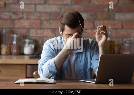 Müder Mann, der eine Brille hält, trockene Augen reibt und am Tisch sitzt Stockfoto