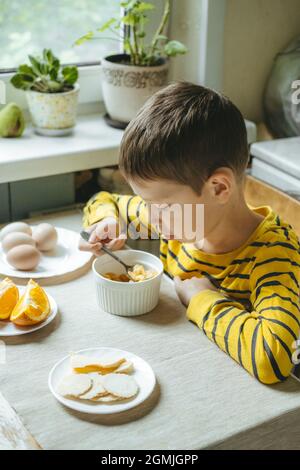 Junge isst morgens Frühstück mit Müsli und Milch. Frühstück in der Küche vor der Schule. Der Junge isst am Tisch am Fenster Stockfoto