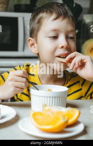 Junge isst morgens Frühstück mit Müsli und Milch. Frühstück in der Küche vor der Schule. Der Junge isst am Tisch am Fenster Stockfoto