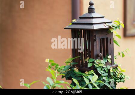 Die Lampe aus Holz hat eine quadratische Form, die einem Haus ähnelt. Installiert entlang eines Gartenweges mit grünen Pflanzen. Stockfoto