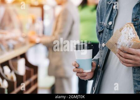 Nahaufnahme eines Bokeh-Bildes von einem Mann in einer Jeansjacke, der eine Tasse Kaffee und ein Croissant in einem Papiertuch hält. Stockfoto