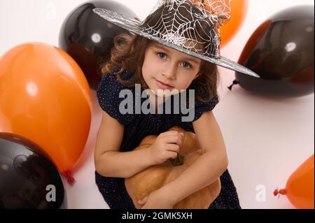 4 Jahre alt schöne hübsche Baby-Mädchen in Hexenkleid und Zauberer Hut sieht Kamera spielen mit Ballons und einem Kürbis isoliert auf weißem Hintergrund. Stockfoto