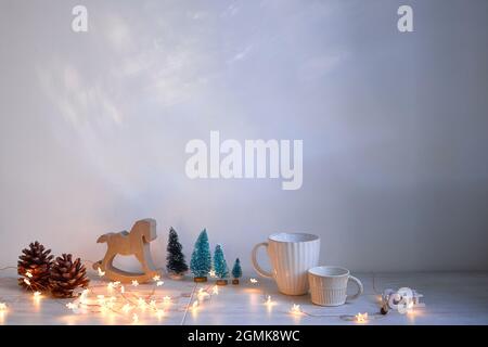 Weihnachtskomposition auf dem Tisch. Graues Schaukelpferd aus Holz, vier kleine künstliche schneebedeckte Bäume, Engel, Girlande und Becher Stockfoto