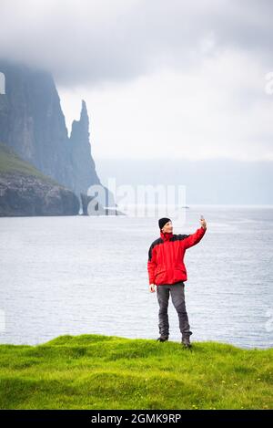 Ein Tourist in einer roten Jacke macht ein Selfie im Hintergrund des Hexen-Finger-Felsens auf den Färöer-Inseln Stockfoto