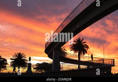 Silhouette bei Sonnenuntergang. St Kilda Melbourne Australien, Silhouetten bei Sonnenuntergang einer Fußgängerbrücke, Palmen und Captain Cook Statue. Stockfoto
