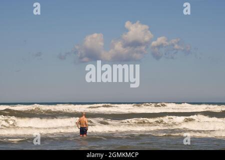 Rückansicht eines Mannes, der an einem windigen Sommertag an der Küste mit hohen weißen Wellen steht, Marina di Castagneto Carducci, Livorno, Toskana, Italien Stockfoto