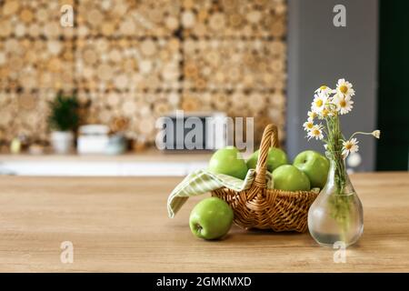 Korb mit frischen grünen Äpfeln und Vase mit Blumen auf dem Tisch in der Küche Stockfoto