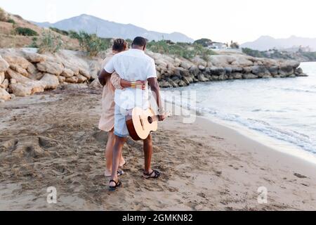Fröhliches gemischtes Rennen, Paar mittleren Alters, das sich umarmt, während man am Strand mit einer Gitarre läuft. Mann mit Gitarre und Frau. Stockfoto