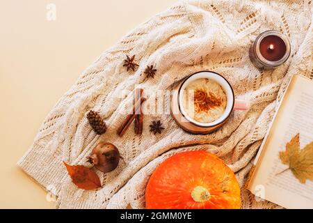 Eine Tasse Kürbis Latte auf dem Tisch mit Buch, Kerze, gelben Blättern und Gewürzen auf einem gestrickten Pulloverhintergrund. Herbstkonzept, Draufsicht, flach liegend Stockfoto