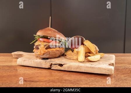 Rinderburger mit geschmolzenem Käse, karamellisierten Zwiebeln, Rucola, Tomaten, Mayonnaise und Senf mit Dorfbrot und Pommes auf Holzbrett Stockfoto