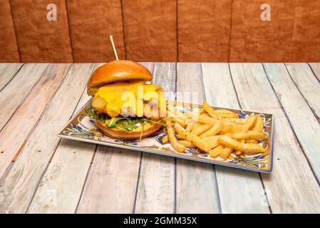 Knuspriger Chicken Burger mit Brioche-Brot, geschmolzenem Cheddar-Käse, eingelegten Gurken, Sesam, Rucola und Senf-Pommes frites Stockfoto