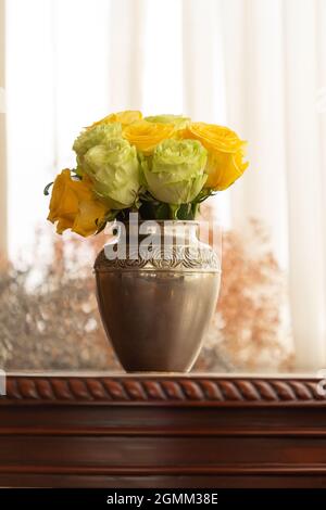 Schöne Metallvase mit einem Blumenarrangement aus gelben und weißen Rosen auf einem Holztisch mit Schnitzerei, dekoratives Objekt im Inneren des Hauses Stockfoto