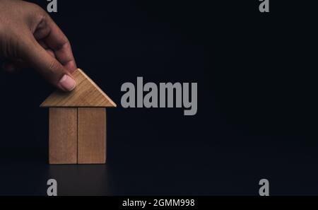 Handbauhaus mit Holzblöcken, Dachverlegung auf dunklem Hintergrund mit Kopierraum. Immobilienplanung. Symbol für den Hausbau, Stockfoto