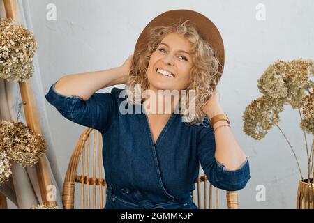 Glücklich lächelnd krauses Blondine in Hut auf hellem Hintergrund zwischen natürlichen Dekor von getrockneten Blumen , Lächeln breit auf die Kamera mit weißem Lächeln. Stock Foto Stockfoto