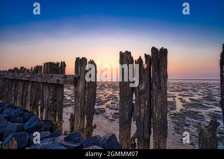 Blick auf das Wattenmeer bei Sonnenuntergang, bei Ebbe. Ein farbenfroher, dramatischer Himmel. Holzpfosten als Silhouette im Schlamm. UNESCO. Wattenmeer-Welterbe Stockfoto