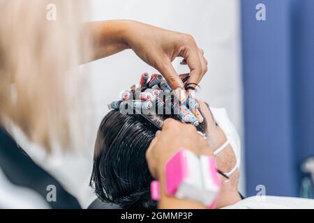 Nahaufnahme der Hände eines Friseurs, der das Haar eines Mannes mit Maske kräuselt Stockfoto