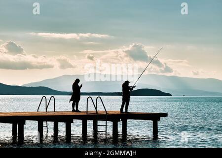 Silhouette einer Frau, die mit einer Angelrute am Pier Fische fängt und ihre Tochter beim Sonnenuntergang ein Foto mit einem Smartphone macht. Stockfoto
