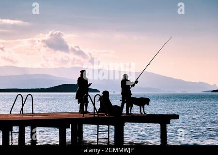 Silhouette einer Frau, die mit einer Angelrute am Pier mit zwei anderen Frauen und einem Hund Fische fängt. Stockfoto