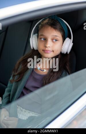 Nettes kleines Mädchen, das Musik in Kopfhörern hört, während es auf dem Rücksitz des Autos sitzt und durch das Fenster schaut Stockfoto