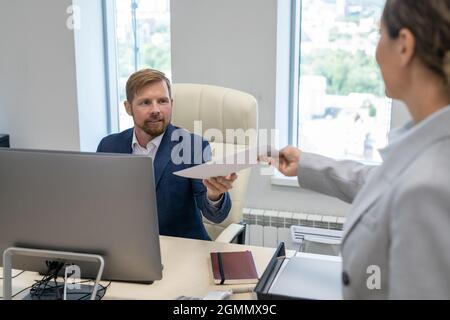 Junger Geschäftsmann, der die von seiner Kollegin oder Sekretärin übergebenen Finanzpapiere nimmt Stockfoto