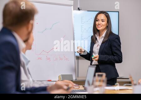 Fröhlicher junger Redner, der vor dem Publikum am Whiteboard steht und einen Graphen zeigt Stockfoto