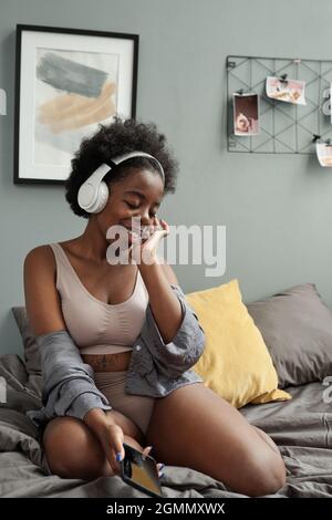 Junge lächelnde Frau, die nach dem Schlafen im Bett sitzt und Musik in Kopfhörern genießt Stockfoto