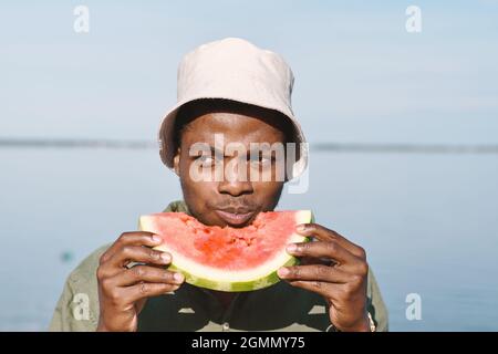 Junger afrikanischer Mann, der am Sommertag am Wasser ein Stück frische Wassermelone isst Stockfoto