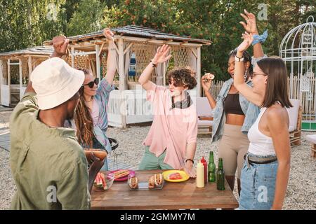 Fröhliche junge Menschen tanzen mit erhobenen Armen am Tisch mit Snacks, während sie eine Party im Freien genießen Stockfoto