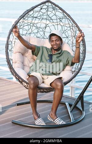 Glücklicher junger afrikanischer Mann in Casualwear, der in einem gemütlichen und bequemen Sessel am See sitzt Stockfoto