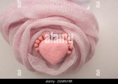 Nahaufnahme der Füße eines Neugeborenen auf einer rosa Decke. Rosa gestricktes Wollherz in den Zehen eines Neugeborenen. Stockfoto