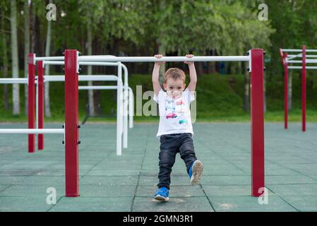 Ein kleiner Junge lernt an einer horizontalen Stange im Freien hochzuziehen. Das Kind hängt an den unebenen Stäben. Stockfoto