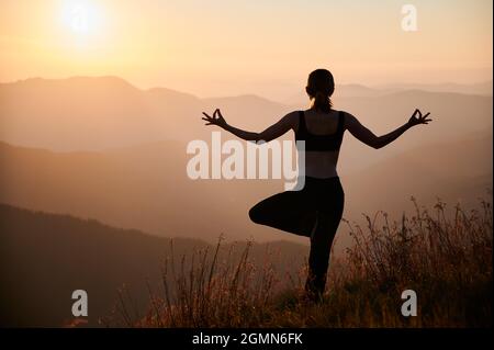 Rückansicht einer jungen Frau, die Yoga-Pose auf einem grasbewachsenen Hügel mit orangefarbenem Himmel auf dem Hintergrund durchführt. Fit Frau steht auf einem Bein und tut Gyan Mudra Handbewegung während Yoga-Übungen im Freien. Stockfoto