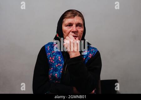 Unschärfe-Porträt der 60er Jahre russische Großmutter ältere alte Frau sitzt drinnen. Alte Frauen im Schal. Traurige, denkende Person aus nächster Nähe. Weißer, schmutziger Hintergrund Stockfoto