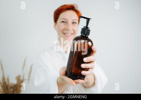 Lächelnde, rothaarige Chemikerin, die eine Shampoo-Flasche mit einer Pumpe präsentiert. Halten Sie es vor der Kamera, und fokussieren Sie das Objekt. Stockfoto