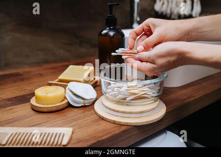 Weibliche Hände, die Baumwollknospen aus einem Glasbehälter aus einem Holzregal aufsammeln. Sie steht vor dem Waschbecken im Badezimmer. Stockfoto